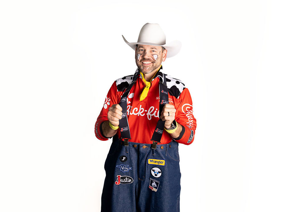 El barrendero profesional Matt Merritt, vestido con una camisa roja, un sombrero de vaquero blanco y sonriendo, sosteniendo las correas de sus tirantes.